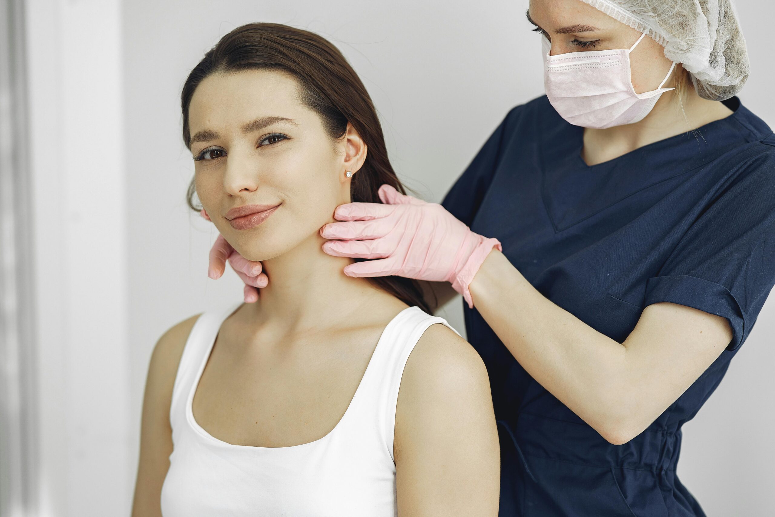 A medical practitioner in scrub suit performs a neck examination on a smiling woman in a clinic.
