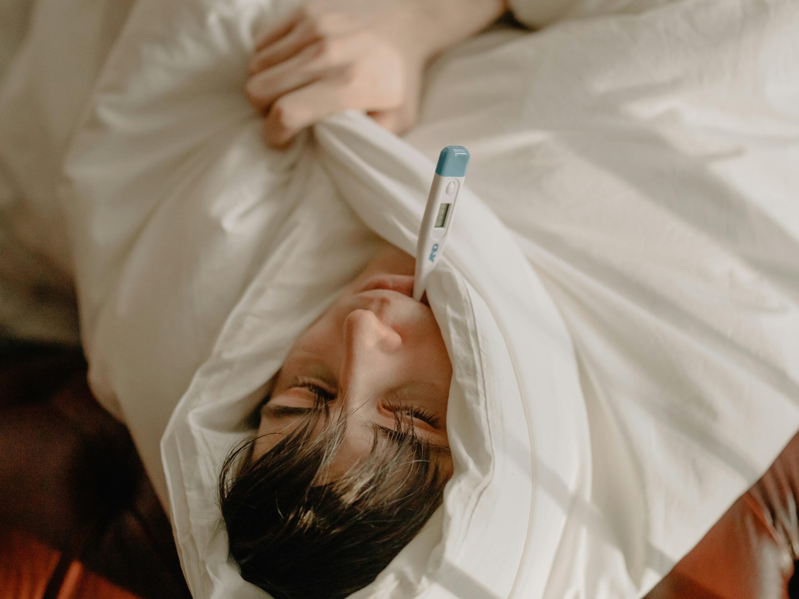 Sick man lying in bed, wrapped in blanket, holding a thermometer in mouth.